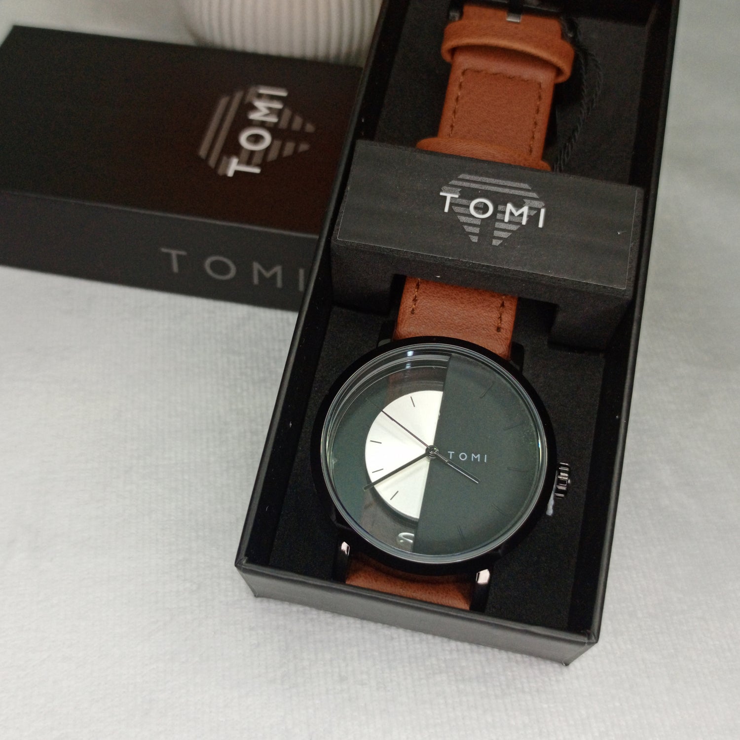 TOMI analog watch with a black dial, brown leather strap, black case, and modern minimalist design.