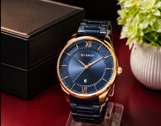 A Curren men's watch with a dark blue dial, gold-tone case, and black link bracelet, displayed on a clear stand next to a black textured watch box and a white vase of green flowers.